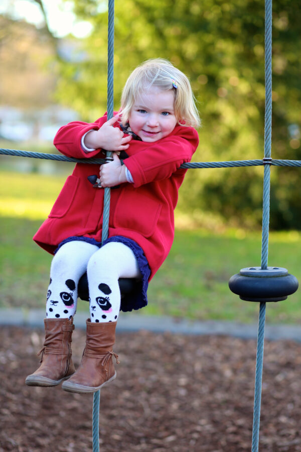 a confident preschooler in a red coat hangs smiling from a climbing net